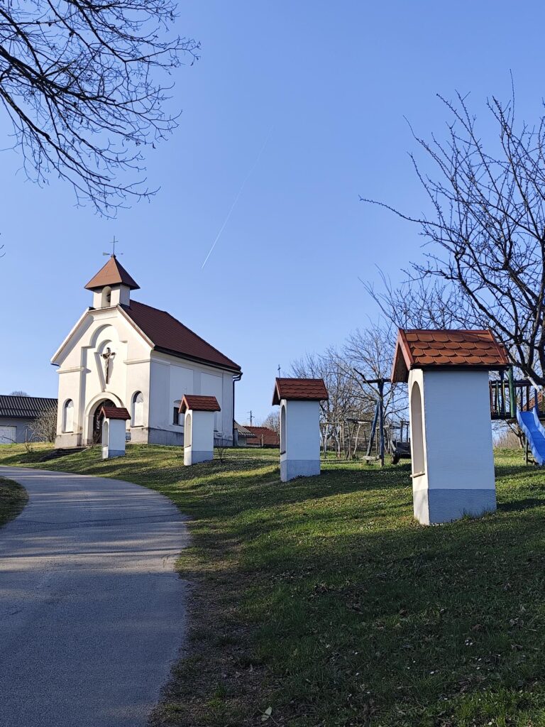 Slika prikazuje cerkev sv. Andreja s štirimi kapelami, ki stojijo ob vaški cesti, ki pelje mimo cerkvice.