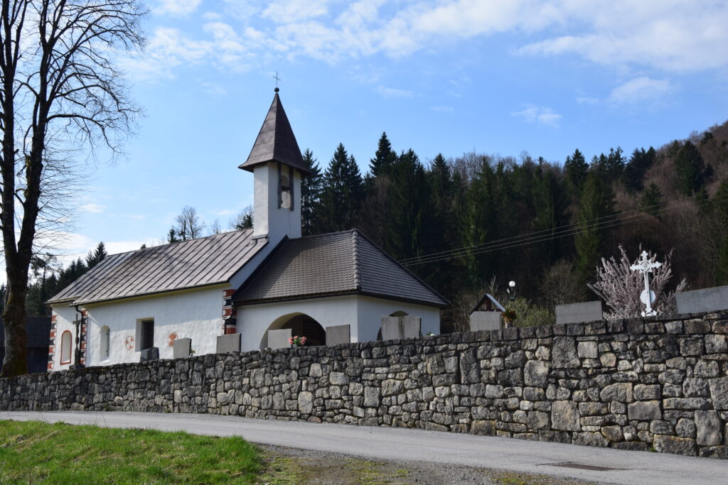 Slika prikazuje cerkev v damlju, ki jo obdaja vaško pokopališče in kamnita škarpa. V ozadju cerkve je gozd.