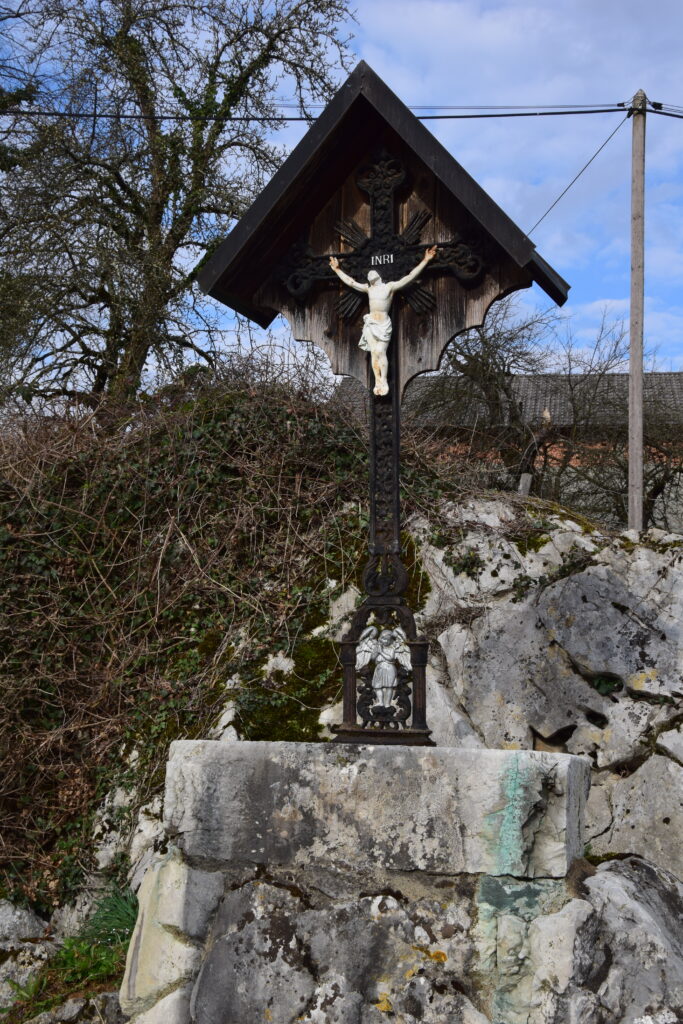 Slika prikazuje cerkveno znamenje na betonskem pomolu,, s križem in križanim Jezusom. Križ je zaščiten s strehico.