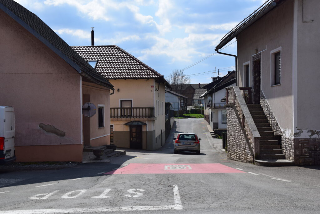 Slika prikazuje trško središče Vinice s stavbami in cesto, ki v križišču vodi proti osnovni šoli in zdravstvenemu domu.